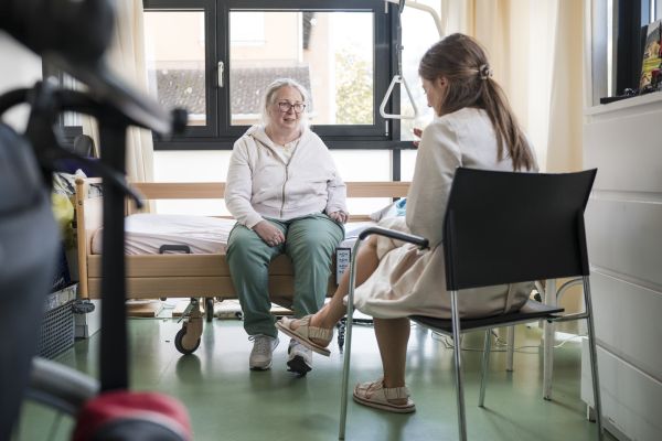 Eine Klientin sitzt auf einem Pflegebett und spricht mit einer Pflegerin
