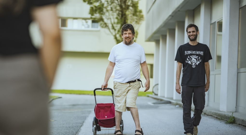 Ein Klient mit seinem Einkaufswagerl geht in Begleitung am Gehsteig zum Einkaufen 