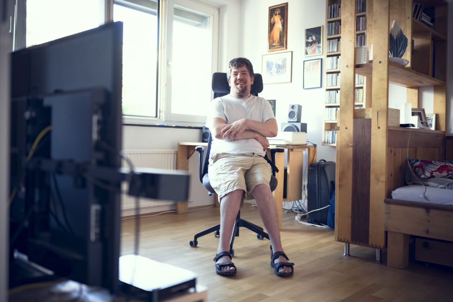 ein Mann mit Behinderung sitzt auf einem Schreibtischstuhl in seinem Zimmer, er lächelt in die Kamera