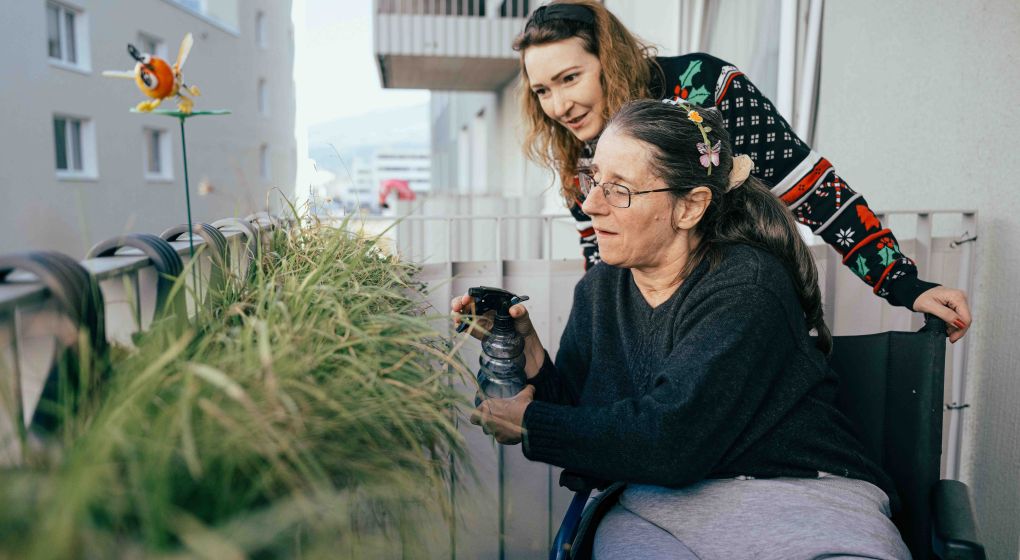 Eine Klientin besprueht zusammen mit ihrer Unterstuetzerin ihre Balkonpflanzen.