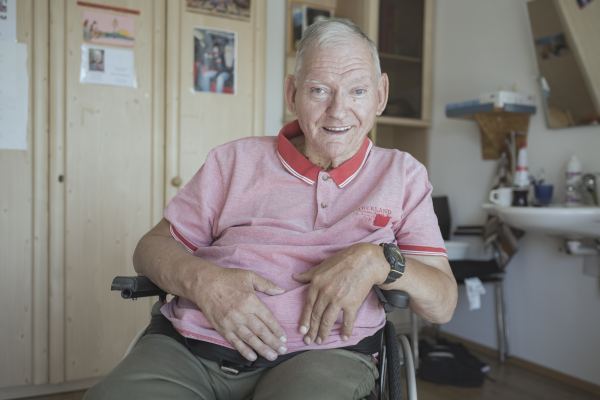 ein Mann mit rotem Shirt sitzt im Rollstuhl; hinter ihm sieht man sein Zimmer; er lächelt in die Kamera