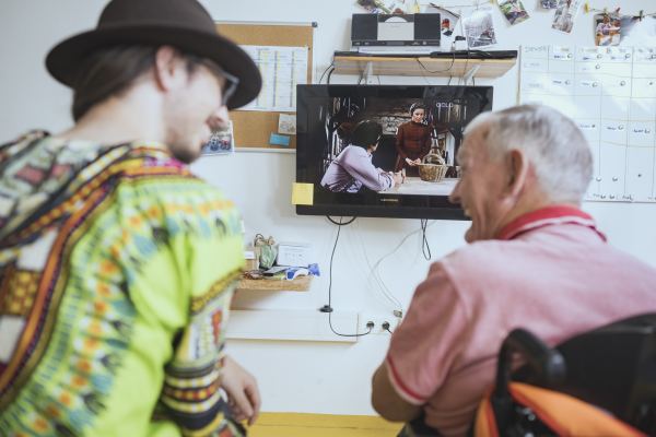 ein Mann im Rollstuhl und ein weiterer Mann mit Hut und buntem Oberteil lächeln sich im Vordergrund an, im Hintergrund läuft ein Fernseher