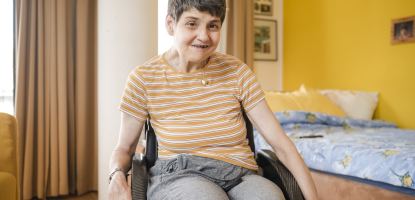 Eine lächelnde Frau in einem Rollstuhl trägt ein gestreiftes T-Shirt und sitzt in einem gemütlichen Zimmer mit gelber Wand und einem Bett im Hintergrund.