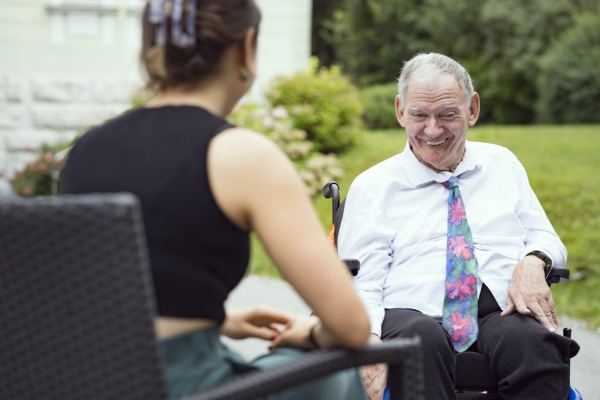 ein Mann im Rollstuhl lächelt; ihm gegenüber sitzt mit dem Rücken zur Kamera eine junge Frau; im Hintergrund sieht man Rasen