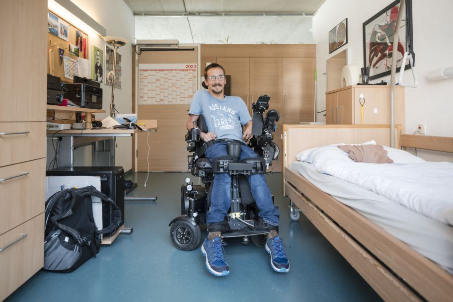 Ein junger Mann mit Behinderung lächelt in die Kamera, er sitzt in einem E-Rollstuhl; um ihn herum sieht man sein Zimmer mit Bett, Schreibtisch und Schrank