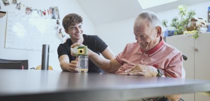 Ein junger Mann hilft  einem älteren, lachenden Mann im Rollstuhl  Saft in ein Glas zu gießen. Sie sitzen an einem Tisch in einem hellen Raum mit einer Pinnwand im Hintergrund.