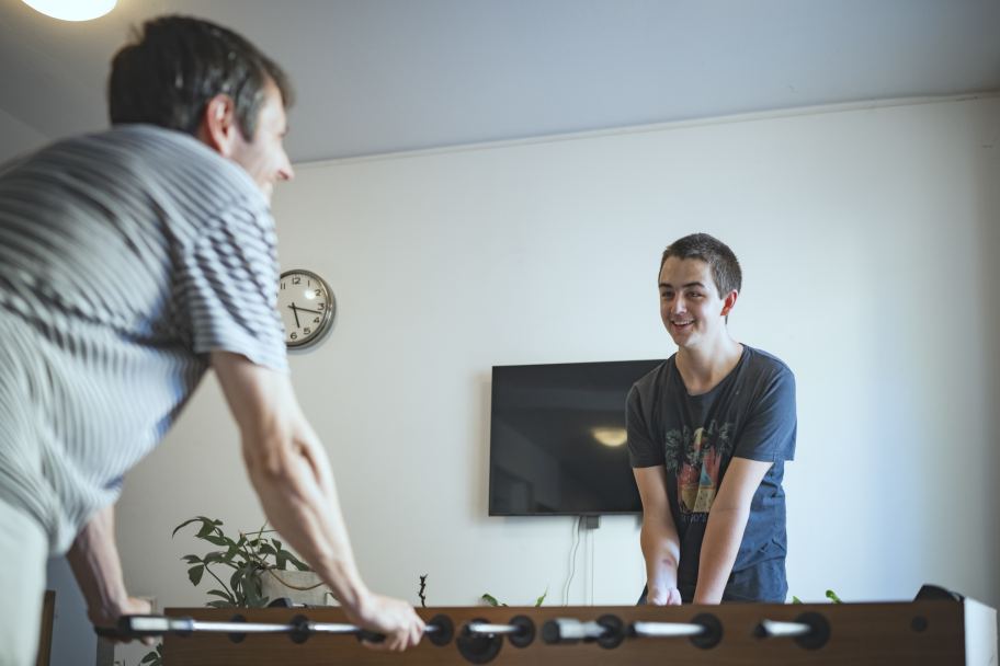 ein junger Mann steht an einem Tischfußball-Tisch; ihm gegenüber steht ein Mitspieler, sie lächeln sich an