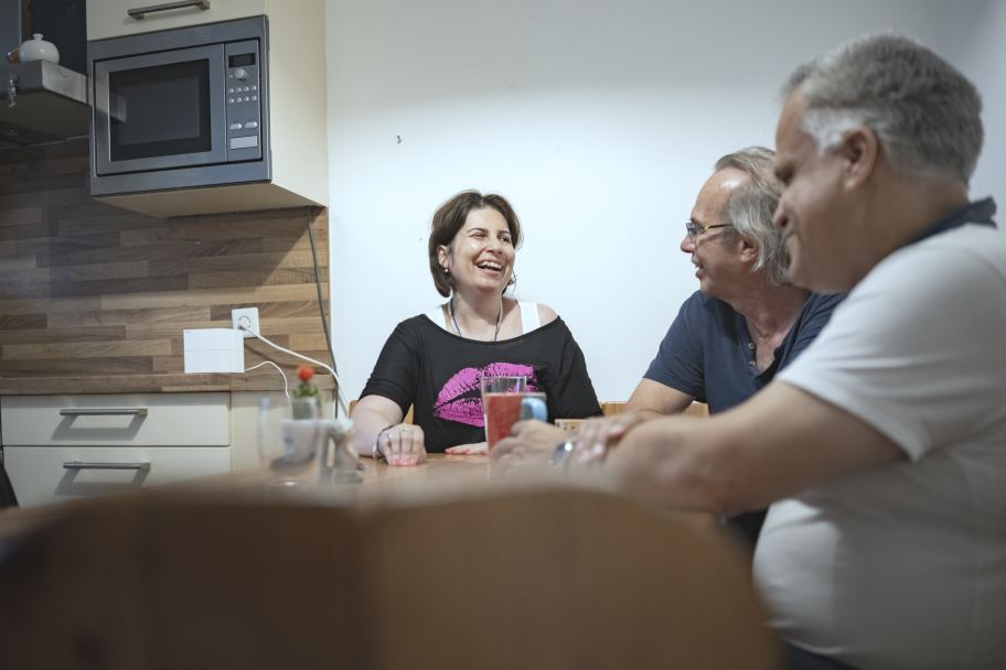 eine Frau und zwei Männer sitzen an einem Küchentisch, quatschen und lachen miteinander